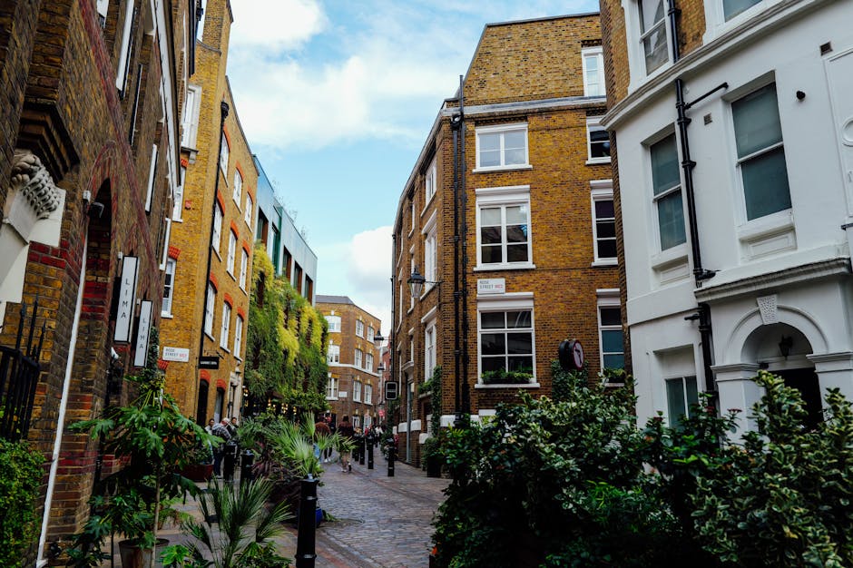A narrow urban street scene viewed from ground level, lined with multi-storey brick and white-painted buildings featuring large sash windows. The street is paved with cobblestones and flanked by black bollards and planters filled with lush green shrubs and small trees. Some buildings have climbing plants growing up their facades, and the sky above is partly cloudy with patches of blue. The scene captures a quiet moment of outdoor city life, with no visible gardening tools or active maintenance but illustrating the potential for garden maintenance and landscaping work in an outdoor residential environment; this image is associated with Gardeners Soho, a company offering gardening services for city gardens and outdoor spaces, illustrating a typical London street that can benefit from professional outdoor care.