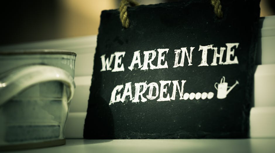 A close-up of a black chalkboard-style sign with white lettering that reads 'We are in the garden', placed on a white wooden surface with horizontal panels. To the left of the sign, a partially visible ceramic or metal container with a smooth, glossy surface and curved shape. The background features blurred horizontal surface details, suggesting a garden or outdoor setting. The scene is lit with soft, natural lighting, highlighting the contrast between the black sign and white text, and emphasizing the textures of the surface and the sign. This image reflects gardening themes associated with outdoor maintenance and landscaping work, relevant to the services offered by Gardeners Soho, especially in the context of garden maintenance and hedge trimming.
